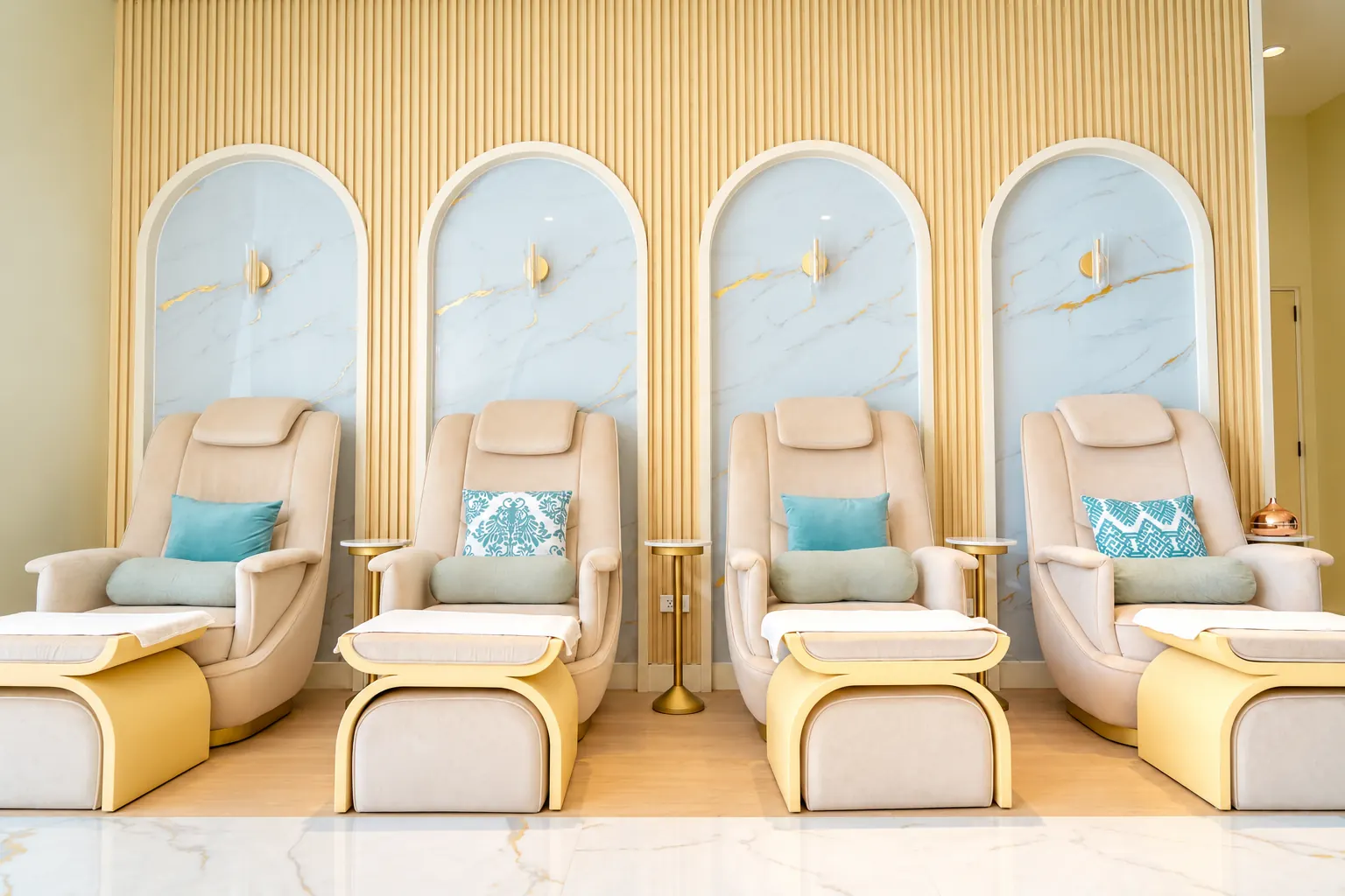 Four cream pedicure chairs with teal cushions, each under a marble arch with a gold sconce, on a pale marble floor